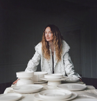A woman with long wavy hair in a textured white jacket stands behind a table displaying the Serax Dune Oval Platter Alabaster -18", surrounded by neatly stacked ceramic plates and bowls. The background is dark and minimal.