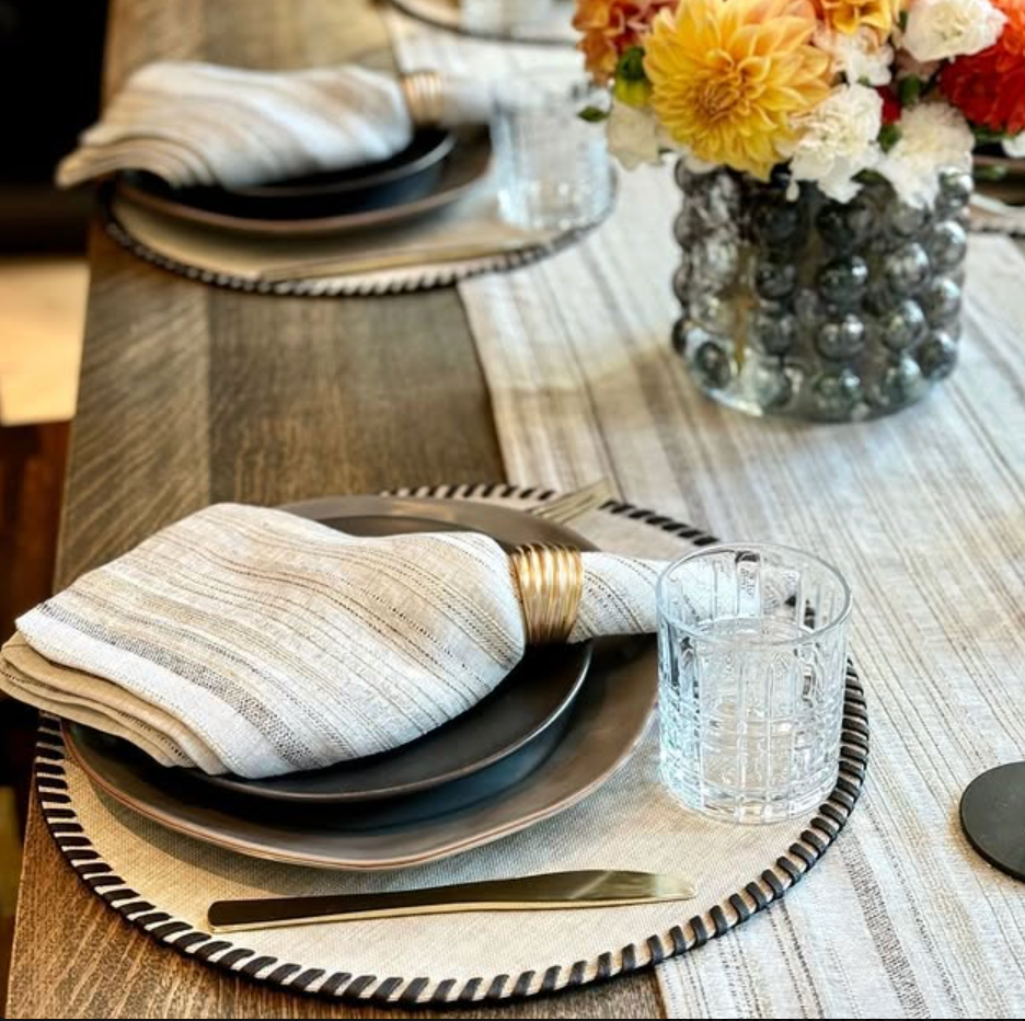 A close-up of a dining table set with gray plates, gold utensils, and Bodrum’s Midas Napkin Set of 4 in gold rings, clear textured glasses, and a centerpiece of colorful flowers in a glass vase.