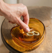 A hand places two gold wedding rings in the heart-shaped Aura Small Catchall Bowl by Yakush, handmade from blown glass, on a wooden surface.