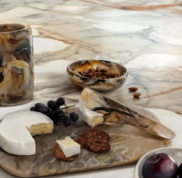 A marble surface displays a cheese platter with brie, crackers, grapes, and plum, styled with almonds and a vase. The elegant setup is complemented by the Nashi Home Arila Cheese Knife - Mocha with its ergonomic handle.