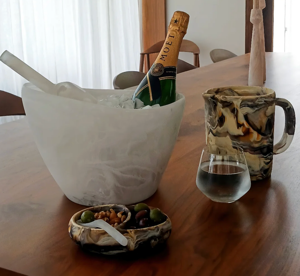 A bottle of Moët champagne in an ice bucket, a marbled pitcher, a glass of water, and the Nashi Home Arila Split Bowl - White Swirl with olives and nuts sit on a wooden table, creating an elegant coastal feel.