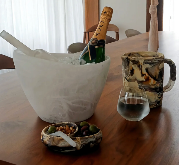 A bottle of Moët champagne in an ice bucket, a marbled pitcher, a glass of water, and the Nashi Home Arila Split Bowl - White Swirl with olives and nuts sit on a wooden table, creating an elegant coastal feel.