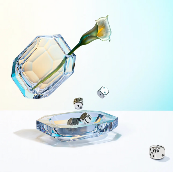 A Baccarat Octogone Bud Vase holds a calla lily, suspended above a matching glass dish. Silver dice scatter and fall midair or rest in the dish, all set against a pastel background reminiscent of Baccarat chandeliers.