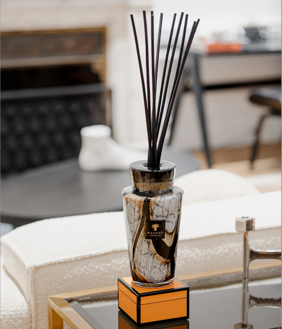A Baobab Stones Diffuser - Marble with black sticks in a hand-blown glass vase sits on an orange box atop a glass coffee table in a modern living room, with a fireplace and blurred furniture in the background for a collector’s display.