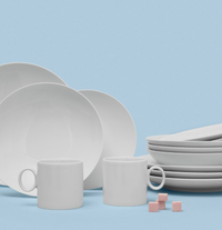 A set of Loft Everyday White Dinnerware by Thomas For Rosenthal, featuring plates and coffee cups, is displayed on a blue background with four pink sugar cubes next to the cups.