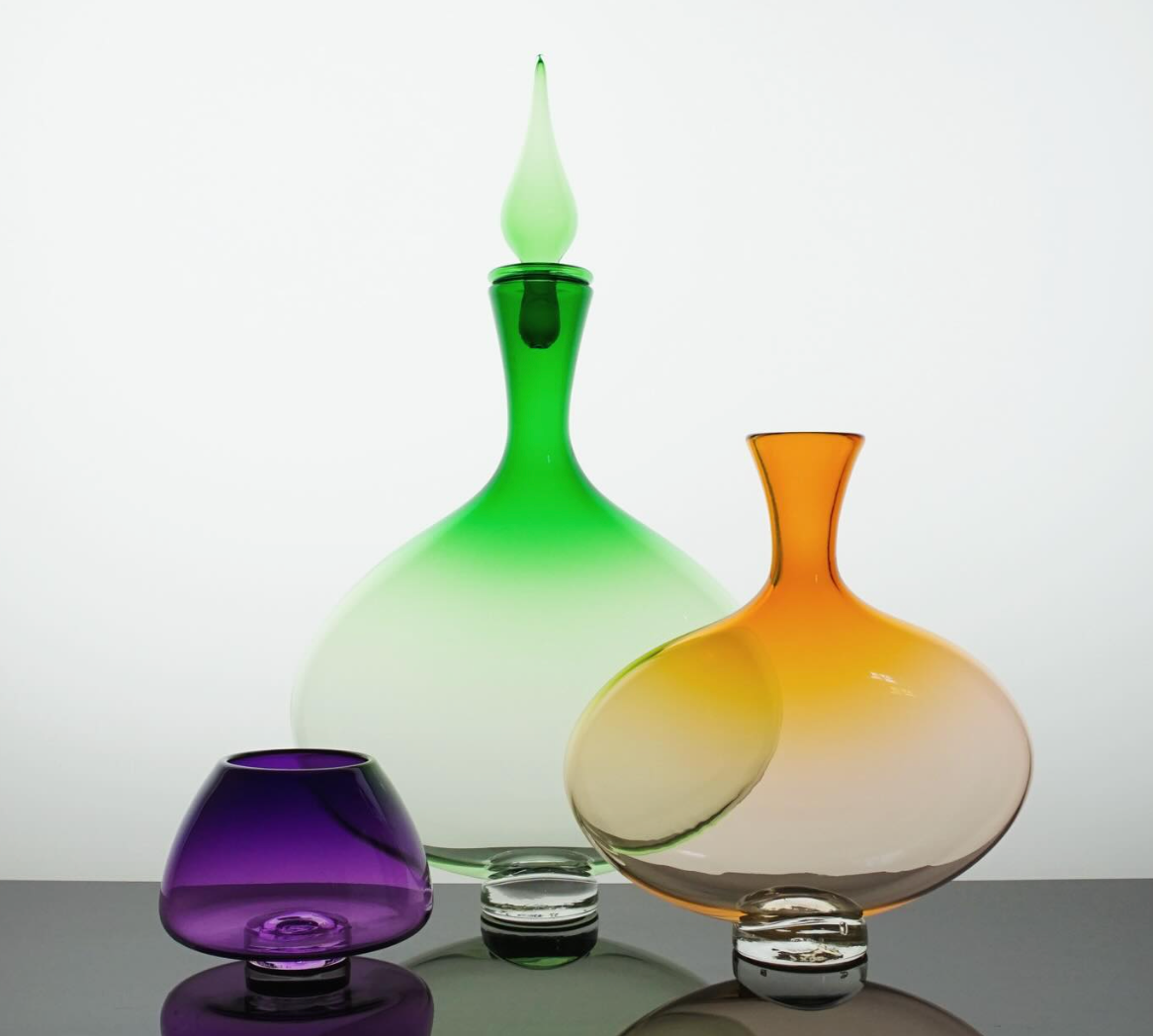 The Andy Koupal Emerald Oval Bottle with Top, along with a round orange vase and a small round purple vase, showcases artisan craftsmanship and clear bases on a reflective surface against a white background.