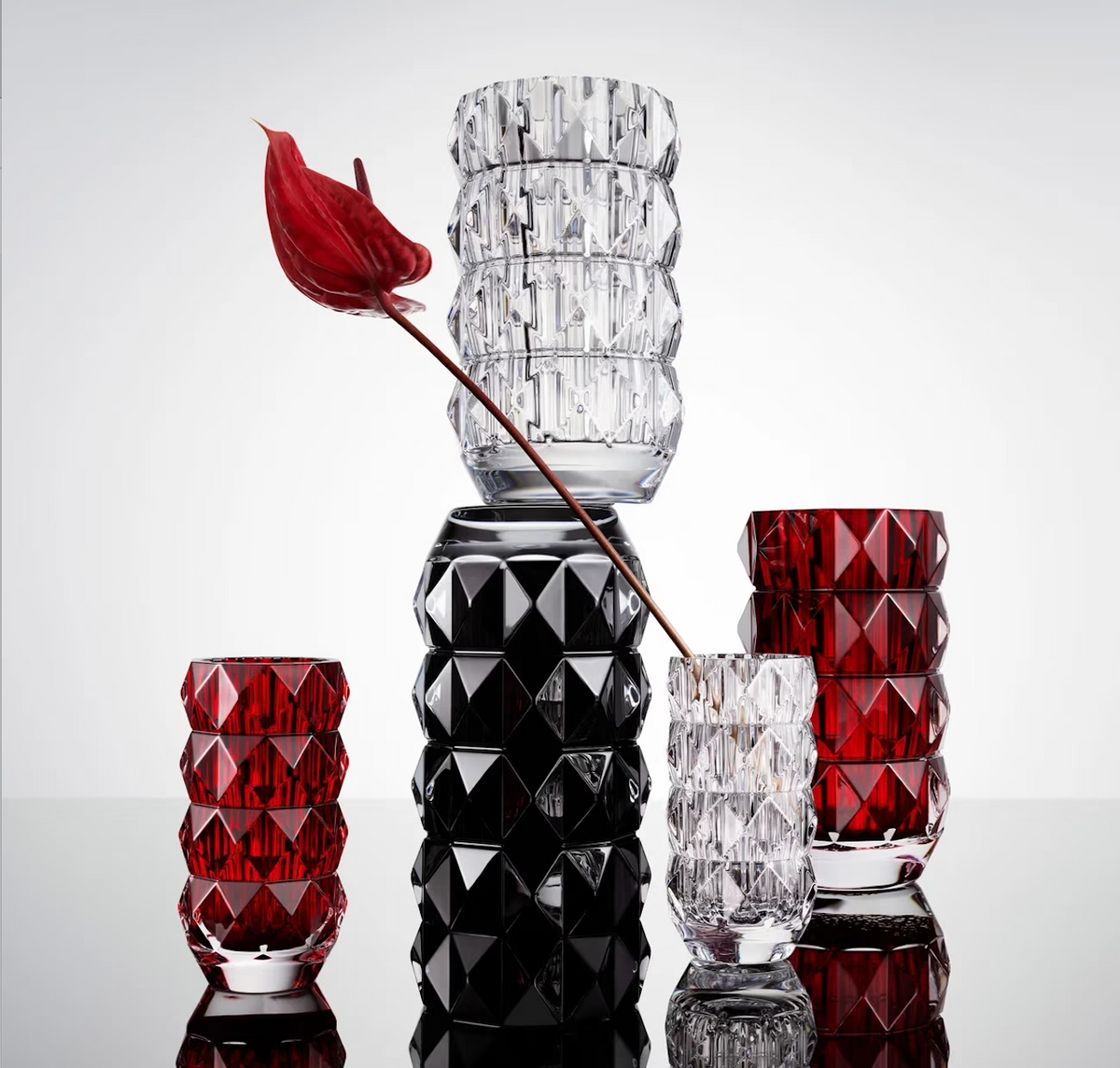 Five geometric glass vases, including the striking Baccarat Louxor Round Vase Red and other elegant crystal vases, stand on a reflective surface; one clear vase displays a single red anthurium with a long stem against a plain white background.