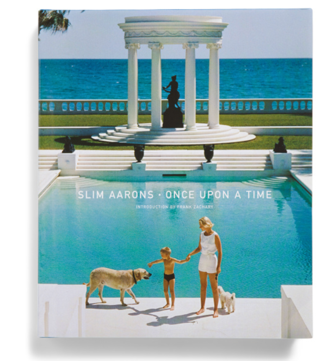 A woman and child with two dogs walk by a pool overlooking the ocean, with a white pavilion and statue in the background—a scene echoing Slim Aarons’ style. The cover reads Slim Aarons: Once Upon A Time (Abrams/Hachette Books).