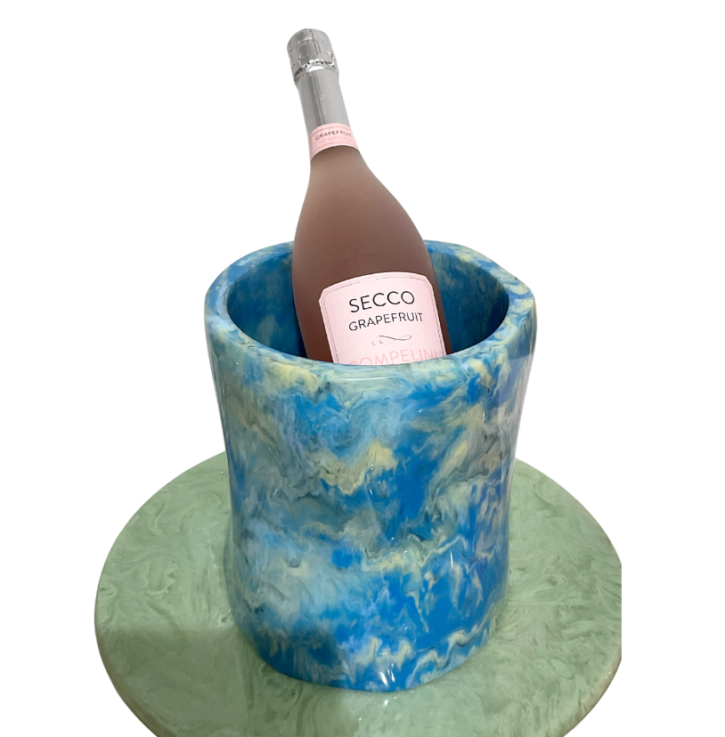 A pink bottle of Secco Grapefruit wine is chilling in a stylish Lily Juliet Resin Ice Bucket, featuring a blue and white marbled design, which is placed on a round, pale green surface.