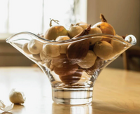 A Simon Pearce Chelsea Bowl, crafted from clear handmade glass, holds assorted onions and garlic on a wooden table, illuminated by soft natural light from a nearby window.