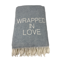 The Wrapped in Love Throw by A Soft Idea is a gray woven blanket with white fringe and "WRAPPED IN LOVE" embroidered on the front. Super soft and machine washable, it makes a thoughtful holiday gift for any cozy home.