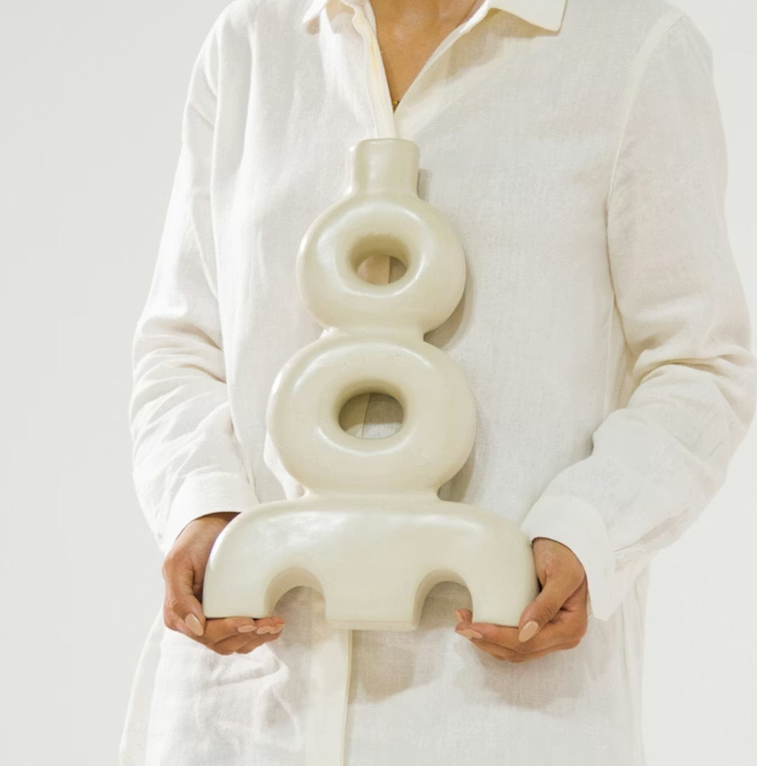 A person in a white button-up shirt holds the Schwung Design Aya Vessel - Eggshell, a modern sculptural vase featuring circular and arch shapes, set against a plain light background.