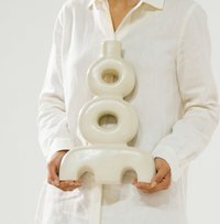 A person in a white button-up shirt holds the Schwung Design Aya Vessel - Eggshell, a modern sculptural vase featuring circular and arch shapes, set against a plain light background.