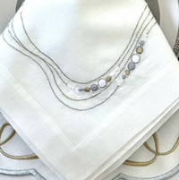 A close-up of Bodrum’s Drift Flow Napkin Gold & Silver Set of 4, crafted from cotton ramie with silver, gold, and grey embroidered curved lines and circular edge patterns.