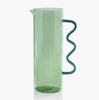 The Zodax Casa Pitcher - Green is a tall, cylindrical glass pitcher with a vibrant green finish and a unique wavy blue handle attached vertically to the side, shown against a plain white background.