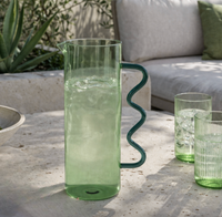 The Casa Pitcher - Green by Zodax, filled with ice water, sits on a stone table beside two matching green glasses, with an outdoor sofa and leafy plants creating a fresh backdrop.