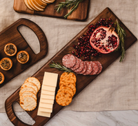 A Bristal Walnut Serving Board by JK Adams holds crackers, cheese slices, salami, halved pomegranate, and rosemary sprigs. Another tray displays crackers and small tarts—all elegantly set out on a neutral tablecloth.