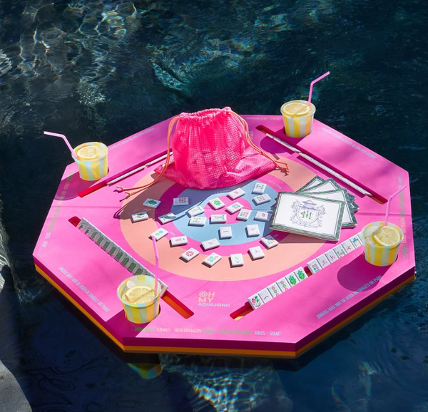 The Oh My Mahjong Mahjong Aqua Pool Mat—a floating pink octagonal table—holds a Mahjong set, four yellow drinks with pink straws, tile racks, and a pink drawstring bag, all resting on the pool’s surface.