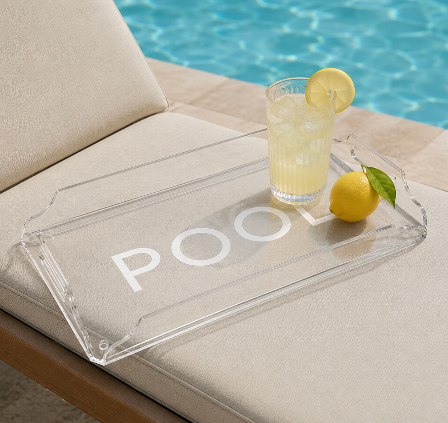 The Eye Candy Deluxe Acrylic Pool Tray, featuring the word POOL, holds a glass of lemonade with a lemon slice and a whole lemon on a beige lounge chair by the pool.