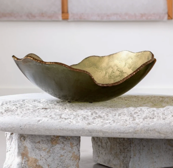 The Mapa Gold & Bronze Trim Bowl by Gardeco, featuring a wavy edge and artisan glass in metallic gold and bronze tones, sits atop a rough, light-colored stone table against a plain wall with framed art partially visible in the background.