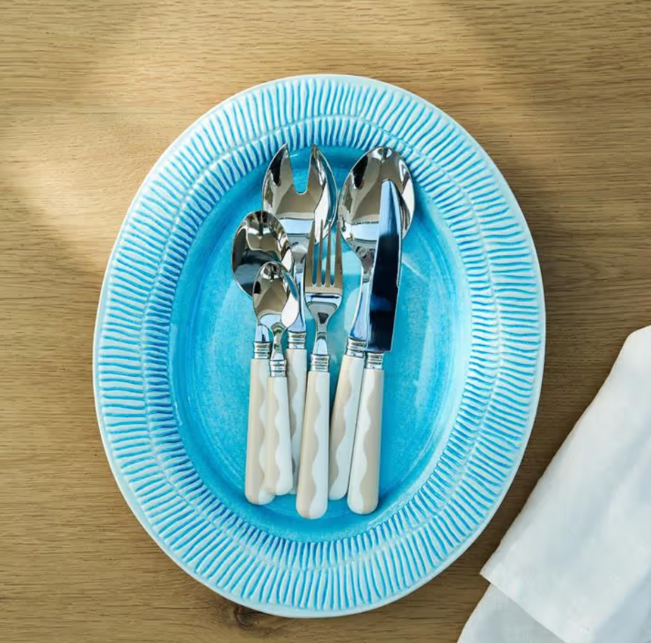 A blue oval plate displays the Addison Ross Wave Outdoor Flatware 4 Sets, featuring elegant silverware with white handles—including a knife, dinner fork, salad fork, soup spoon, and dessert spoon—crafted from 18/10 stainless steel. The plate is arranged on a wooden table beside a folded white napkin.