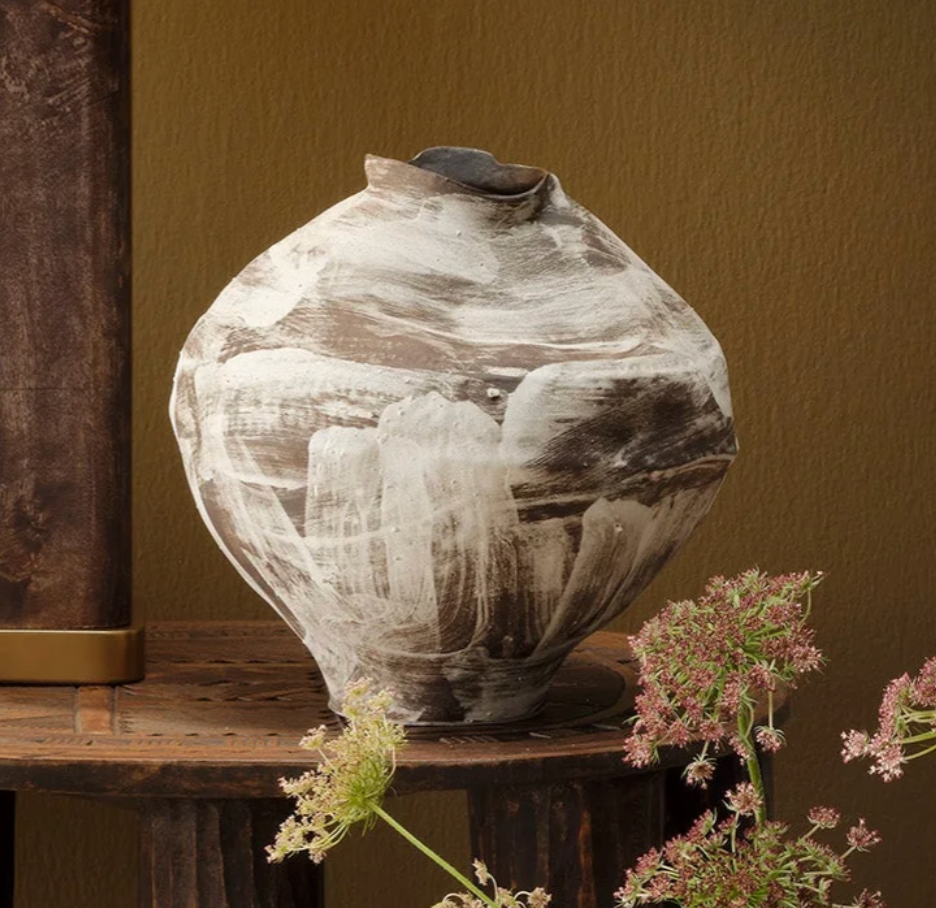 The Moritz Vase - Brown by Jamie Young, featuring a handcrafted ceramic design with a round, asymmetrical silhouette and abstract white and brown brush strokes, sits atop a rustic wooden table. Delicate flowers in the foreground contrast against a tan background, while the modern neutral tones of the vase evoke understated elegance inspired by Italian antique style.