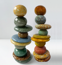 Ten vibrantly colored, rounded, and glossy ceramic forms are artfully balanced in the Axiom Pohacu Sculpture 10 Stacked Stones 26″, displayed on a white surface against a plain white background. Each piece features uniquely sized and patterned discs, spheres, and ovals in both earthy and vivid hues evoking a serene harmony reminiscent of Robert Madvin’s signature style.