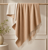 A Thread & Butter Reversible Alpaca Throw in Beige & Ivory hangs neatly over a wooden rod, while a beige vase filled with green leafy branches sits nearby, all set against a neutral paneled wall background.
