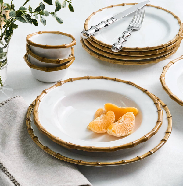 Juliska Bamboo Dinnerware, featuring white plates with bamboo-style rims—including a dinner plate and matching cake plate—are artfully arranged on the table. Some plates are stacked with forks and knives, while one holds vibrant orange slices. Coordinating small bowls from the Juliska Bamboo Dinnerware line are neatly stacked nearby, and a cloth napkin along with green foliage complete the inviting tablescape.