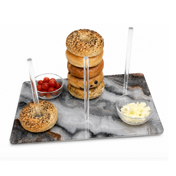 The Continental Bagel Board - Grey Marble Slice by Eye Candy offers an elegant way to serve bagels, featuring a sleek grey marble design with three clear rods for stacking bagels and two small glass bowls—one holding cherry tomatoes and the other filled with crumbled cheese.