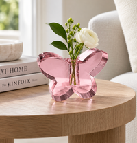 The Crystal Butterfly Bud Vase - Pink by Tizo, featuring a delicate butterfly design and filled with green leaves and white flowers, sits on a round wooden table beside stacked books and a white bowl.