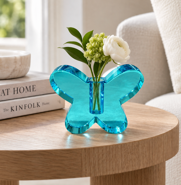 A Crystal Butterfly Bud Vase - Sea Blue by Tizo, filled with green leaves and white flowers, rests on a round wooden table near books and a softly lit window.