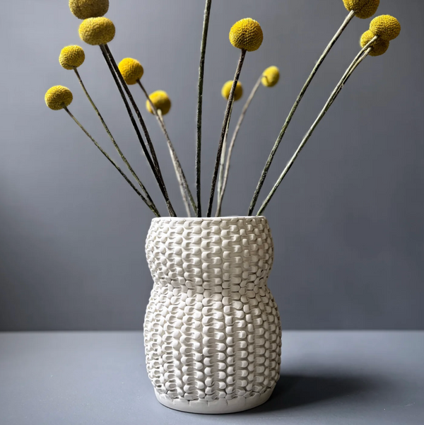A cream-colored Chain Curve Vase from CYM Warkov Ceramics features a textured, woven pattern and holds long stems with round yellow billy ball flowers, creating a striking decorative accent against a plain gray background.