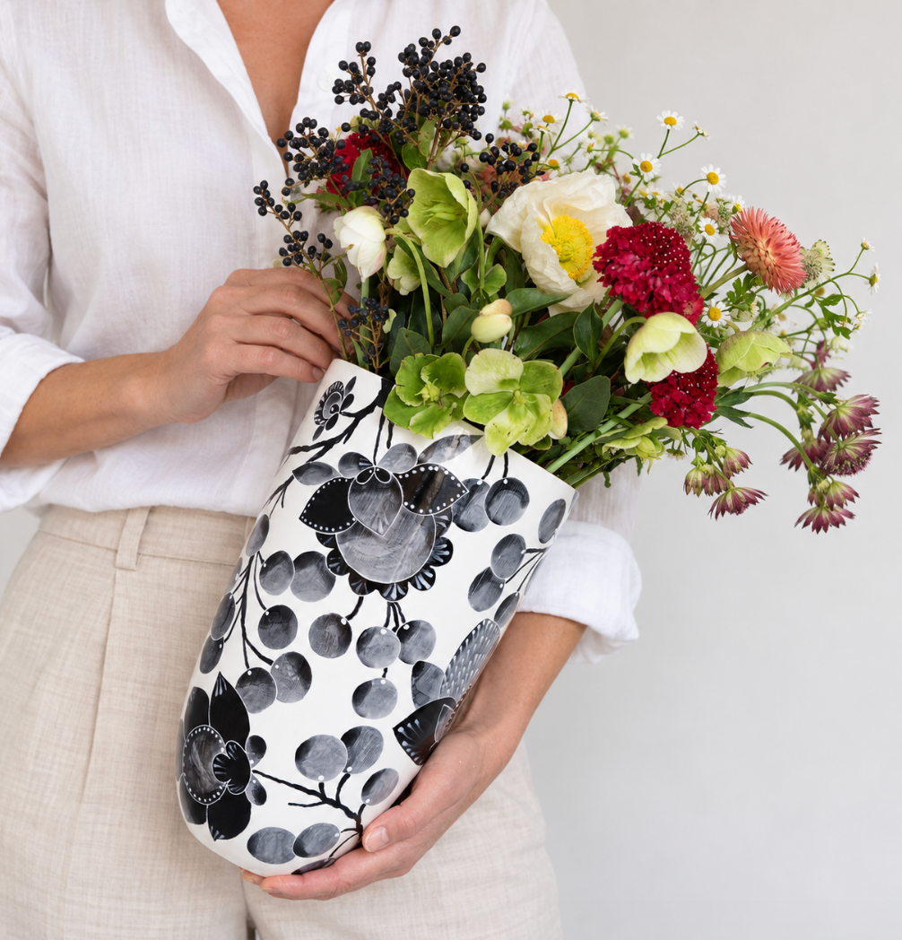 A person in a white shirt and beige pants holds a Jill Rosenwald Studio Bullet Vase - Bramble - Black, filled with colorful fresh flowers, against a plain background.
