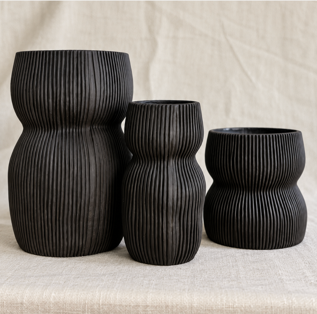 Three CYM Warkov Ceramics Curvy Oval Vases in black, each featuring ribbed ceramic textures and elegantly varied heights, are arranged on a beige fabric surface against a textured, neutral backdrop—embodying the sophisticated charm of artisan craftsmanship.