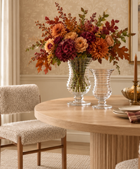 A round wooden dining table with textured chairs features a large glass vase displaying an intricate arrangement of autumn blooms, the Ripples Footed Vase by William Yeoward standing empty beside it, a gold candlestick, and a bowl of small pumpkins—all beautifully set in a warmly lit, elegant room.