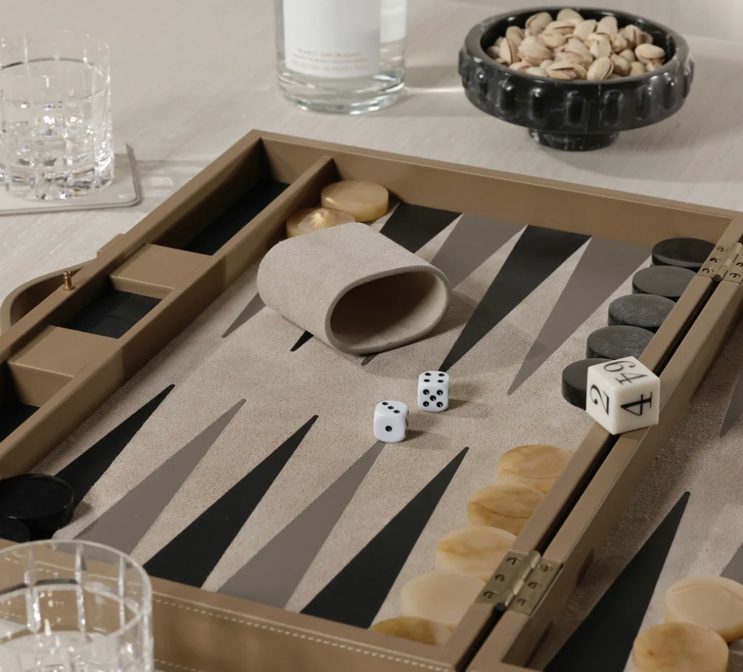 The Gene Leather Backgammon - Taupe by Zodax, a luxurious handcrafted game set complete with dice, checkers, and a dice cup, rests elegantly on a light-colored table. Nearby, glasses of water, a bowl of pistachios, and a bottle enhance the sophisticated atmosphere—perfect for an upscale gaming experience.