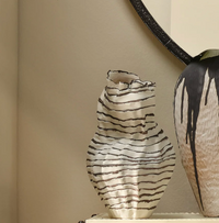 The Cocoon Vase - 15.5″ by Jamie Young, translucent white with black stripes, sits beside a larger round reactive glaze vase with black drips, both reflected in a round mirror against a beige wall.