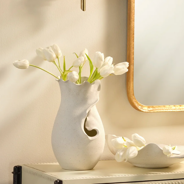 White tulips in the Jamie Young Fabio Vase sit on a cream table next to a matching bowl with tulip petals, beside a gold-framed mirror on a light wall.