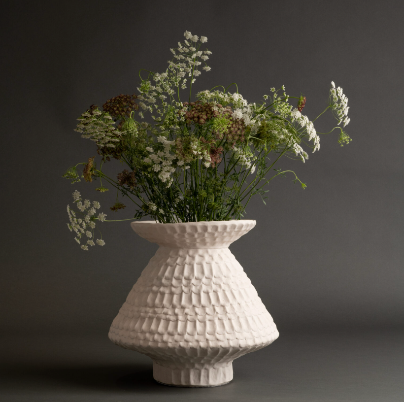 The Teramo Vase by FlowDecor Inc. is a white ceramic piece with a scalloped texture, wide base, and narrow opening, shown filled with white wildflowers and green stems against a dark gray background.