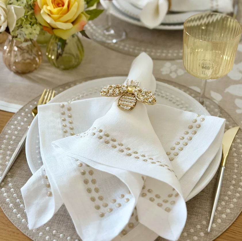 A white linen napkin from the Pearls Napkin Set of 4 - Beige, decorated with a beige beaded border and held by a jeweled bee napkin ring on a white plate.