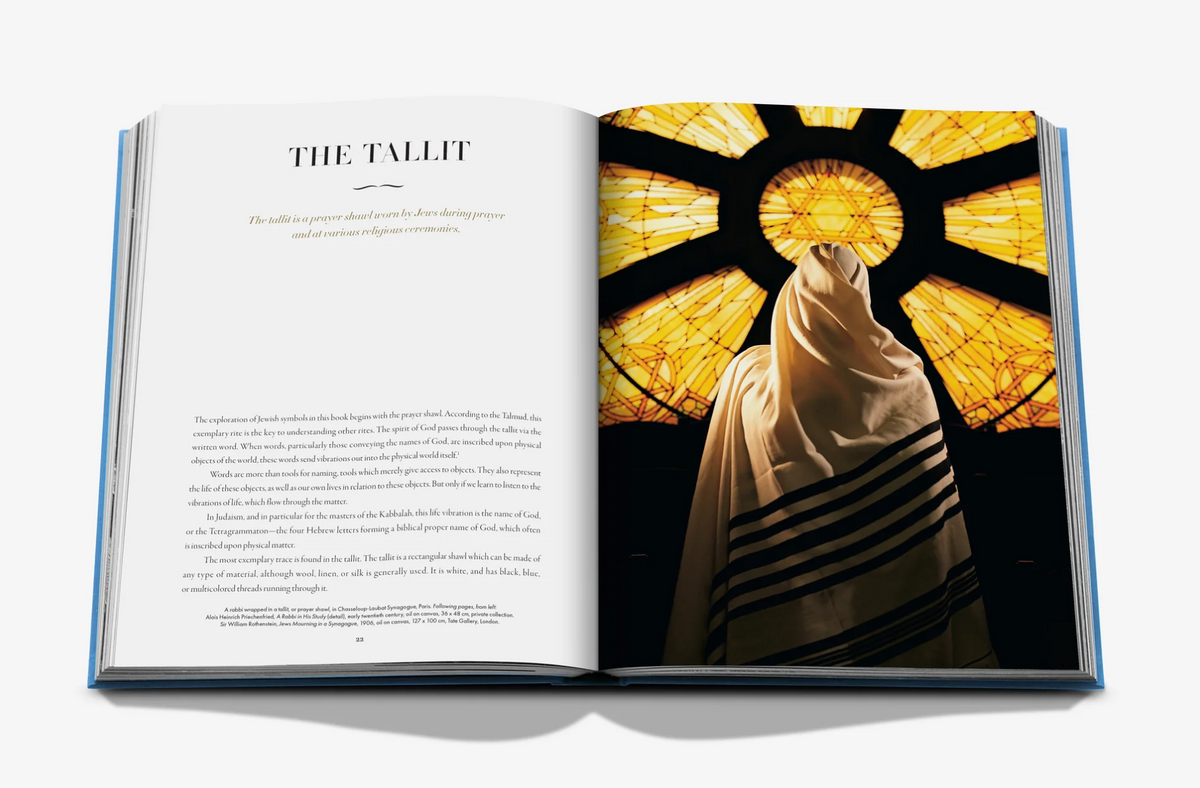 An open book titled Symbols of Judaism displays a page about the tallit next to a person viewed from behind wearing the traditional prayer shawl, standing before a glowing stained-glass window.