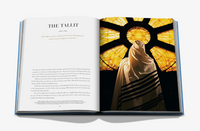 An open book titled Symbols of Judaism displays a page about the tallit next to a person viewed from behind wearing the traditional prayer shawl, standing before a glowing stained-glass window.
