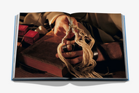 A person wearing tefillin on their hand and arm while holding the tzitzit of a tallit over a book with two tefillin boxes nearby, as seen in Symbols of Judaism by Assouline.