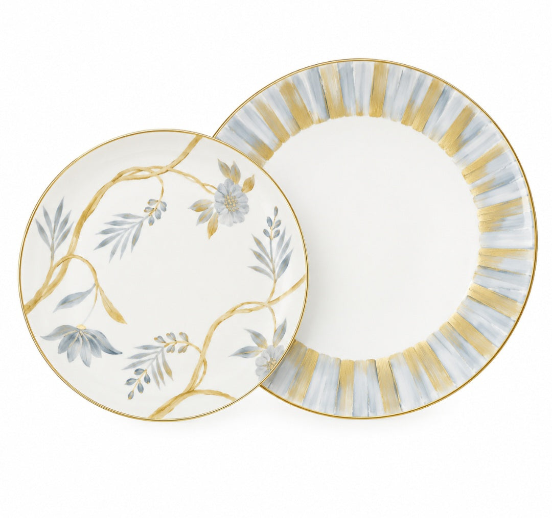 Two Marie Daâge Bouquet De Vie 1 dinnerware plates, one with hand-painted blue flowers and vines, the other with a gold and blue striped border, both featuring gold rims.