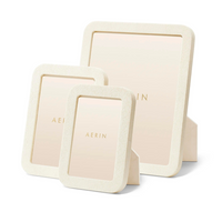 Three Modern Shagreen Frame Creams by Aerin, in various sizes, are shown upright with easel backs. Each features an ivory embossed texture and a beige insert with "AERIN" printed in gold at the center.