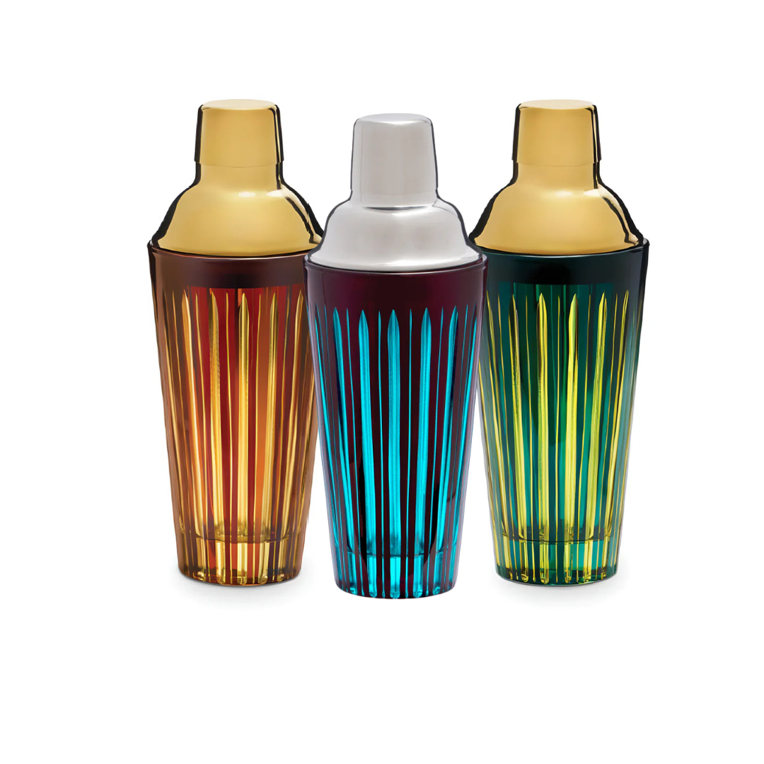 The L'Objet Prism Cocktail Shaker collection features three hand-crafted glass shakers in amber, blue, and green with vertical stripes and gold or silver-tone lids, displayed upright on a white background.