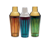 The L'Objet Prism Cocktail Shaker collection features three hand-crafted glass shakers in amber, blue, and green with vertical stripes and gold or silver-tone lids, displayed upright on a white background.