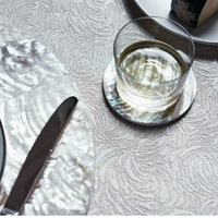 A glass of water sits on a Von Gern Home Shell Coaster Set & Holder White Pearl, with cheese and a knife on matching coasters, all arranged on a textured patterned tablecloth.