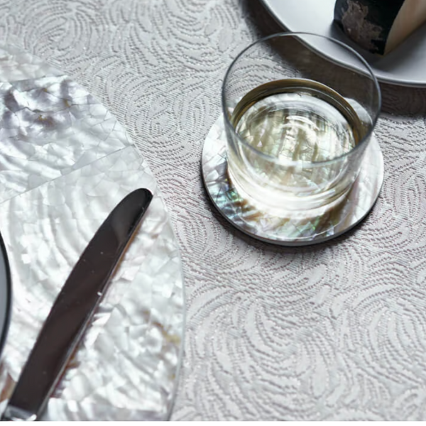 A glass of water sits on a Von Gern Home Shell Coaster Set & Holder White Pearl, with cheese and a knife on matching coasters, all arranged on a textured patterned tablecloth.